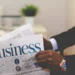 A man in a smart suit reads a business newspaper, with stocks and shares printed on the front cover.