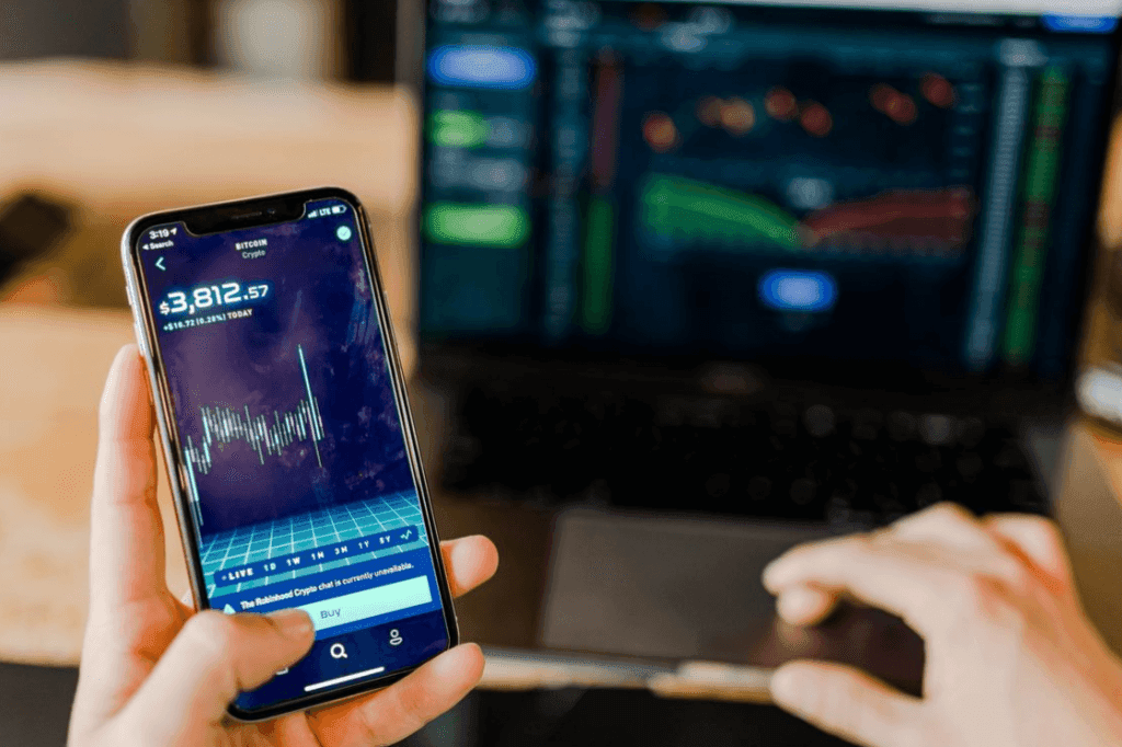 A white hand holds a cell phone demonstrating a fluctuating value of cryptocurrency and sits in front of a computer screen demonstrating a similar trajectory.