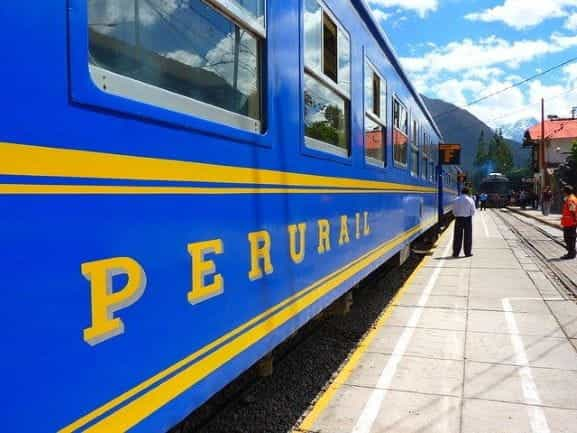 A blue Perurail train at an outdoor train station on a sunny day.