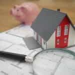 A magnifying glass next to a model of a house and a piggy bank.