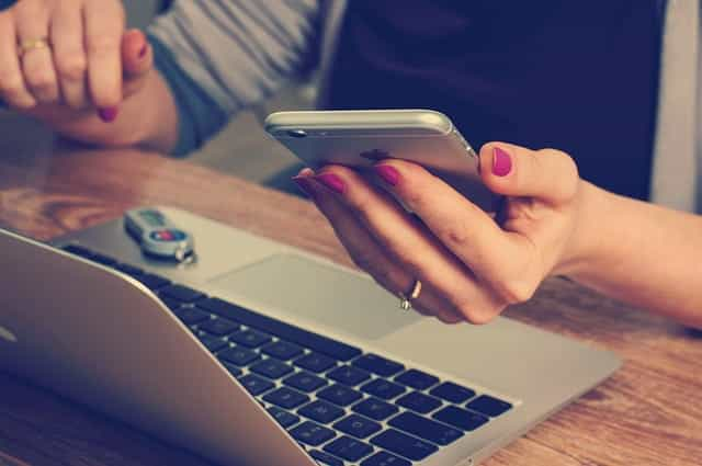 A woman sitting in front of her laptop at a desk while holding a smartphone in her left hand.