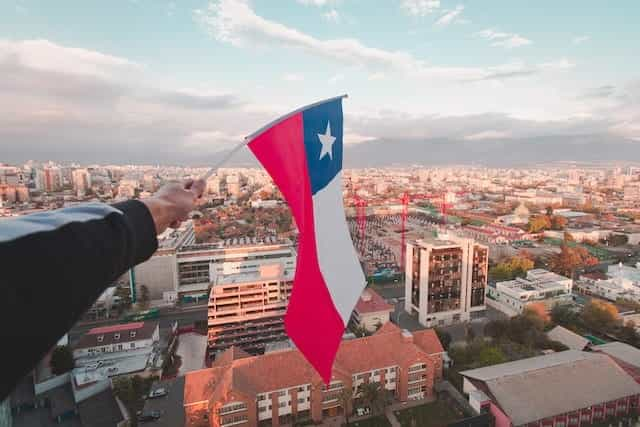 A hand holds the Chilean flag over the city of Santiago from a great height.