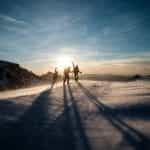 Three people with skiing gear walk towards the summit of a snow covered mountain as the sun sets.