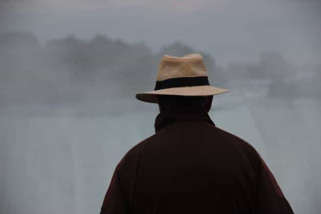 The back of a man wearing a brown jacket and a beige fedora and looking into the mist.