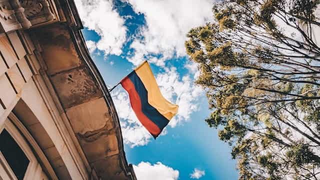 The Colombian flag waves between a building, trees, and a blue sky.