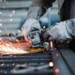 A worker using a blade saw on a piece of metal in a factory, with sparks flying out from the blade grinding against the metal.