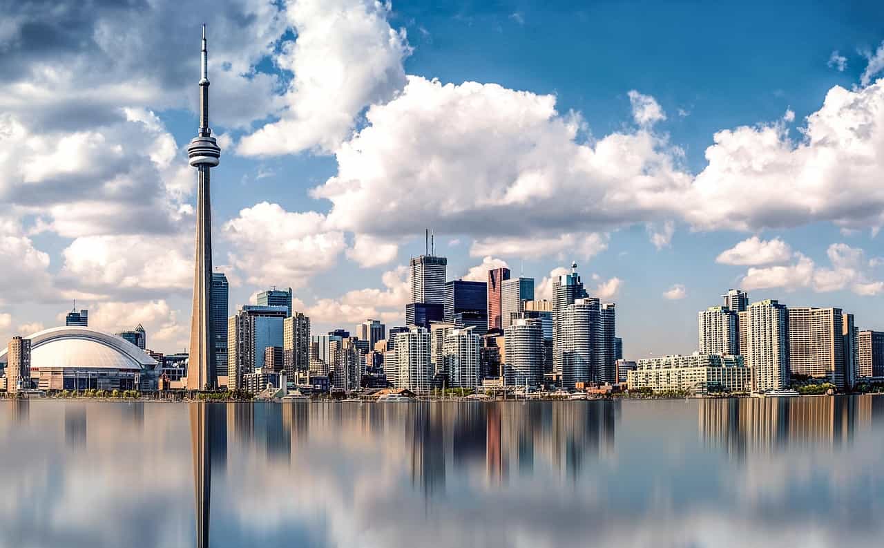 The skyline of downtown Toronto, Ontario during the day, featuring several tall buildings and skyscrapers, as well as the world-famous CN Tower, all of which are reflecting on the surface of Lake Ontario in the foreground.