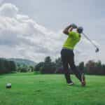 A male golfer post-golf swing on an outdoor golf course, with rolling green mountains seen off in the distance.