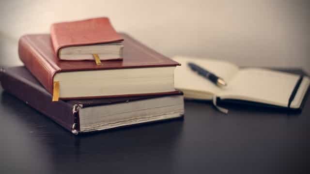 Three heavy books stacked on top one another on a desk next to an open notebook with a pen on top.